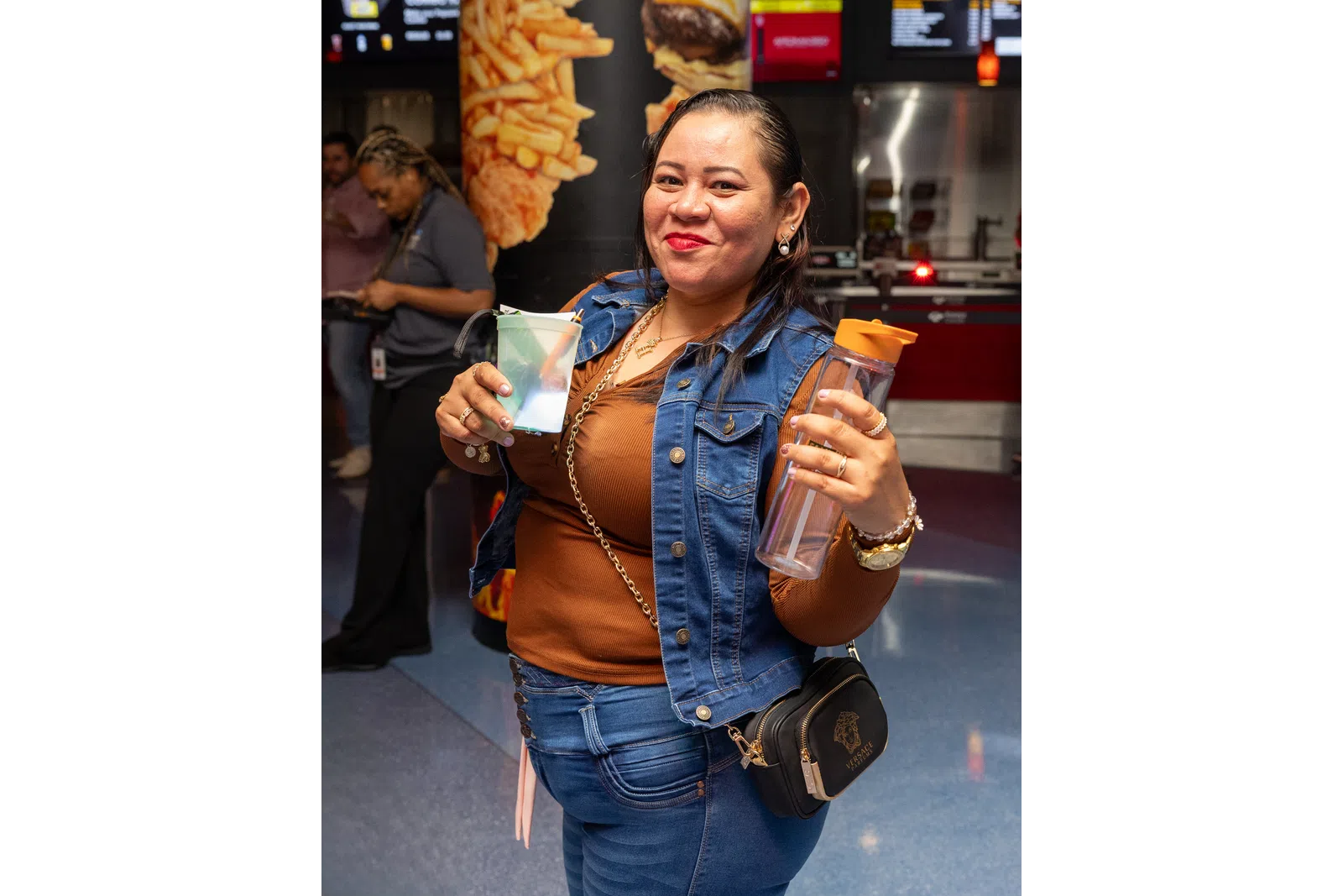 Smiling woman holding a clear cup and water bottle in a casual denim outfit at a fast food restaurant.