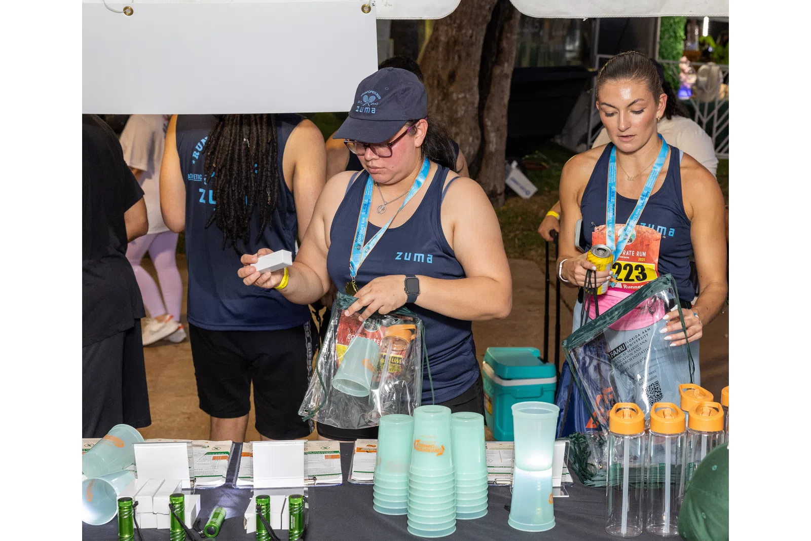 Two women wearing Zuma shirts distributing cups and water bottles at an outdoor event booth.