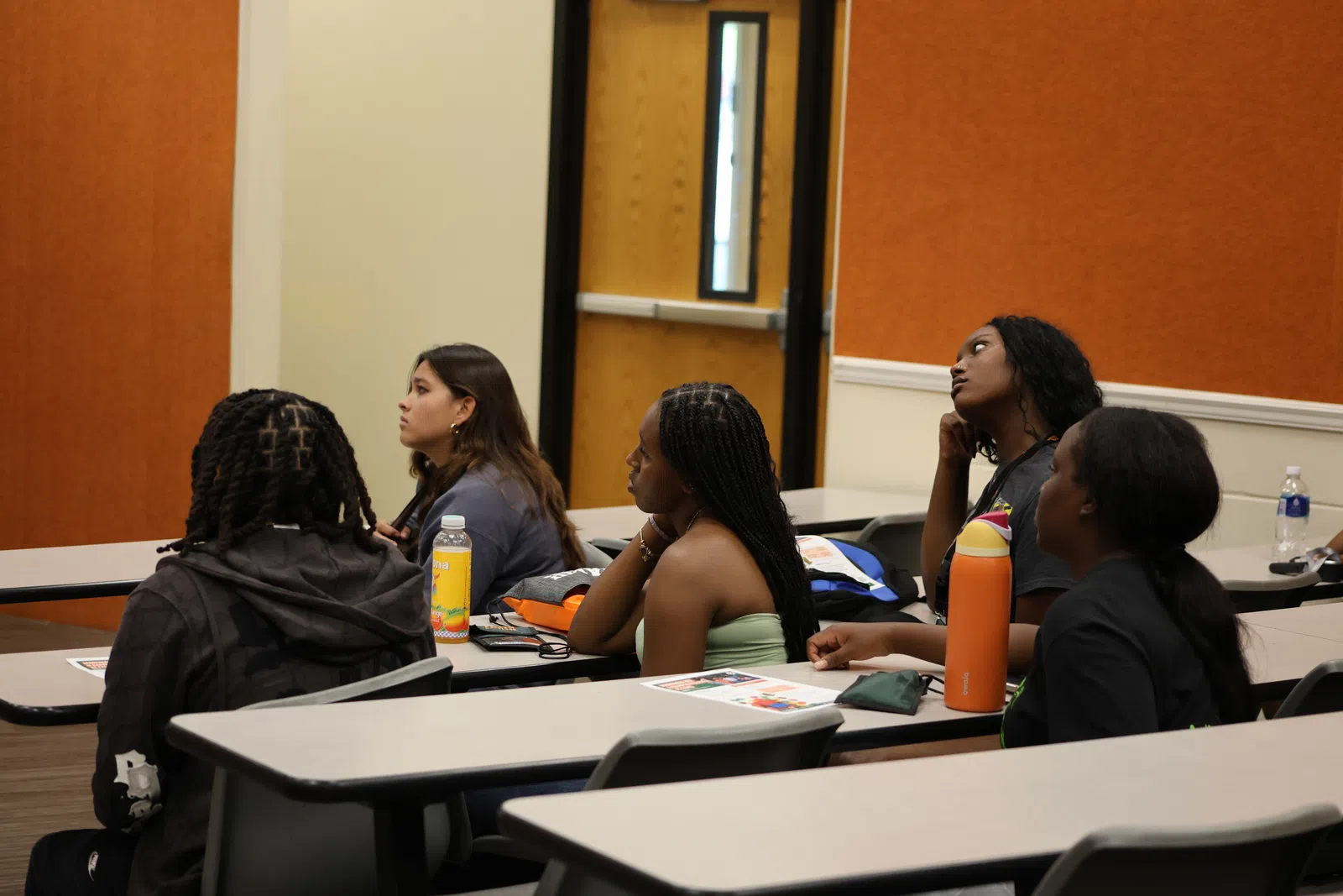 Students attentively listening during a lecture in a classroom with orange walls and modern desks.