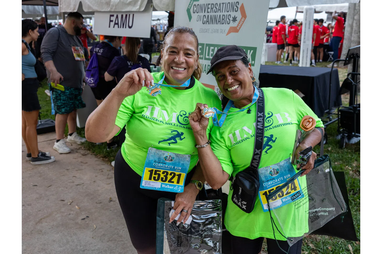 Two women in bright green run shirts proudly display medals at a community race event with FAMU sign visible.
