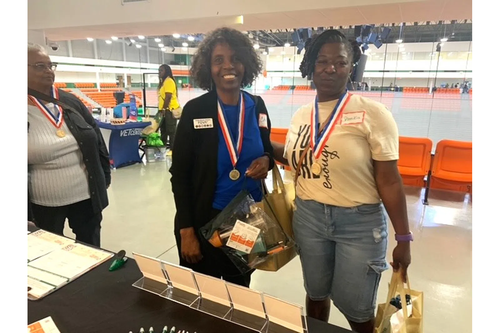 Two women wearing medals holding gift bags at an indoor event with orange seating in the background