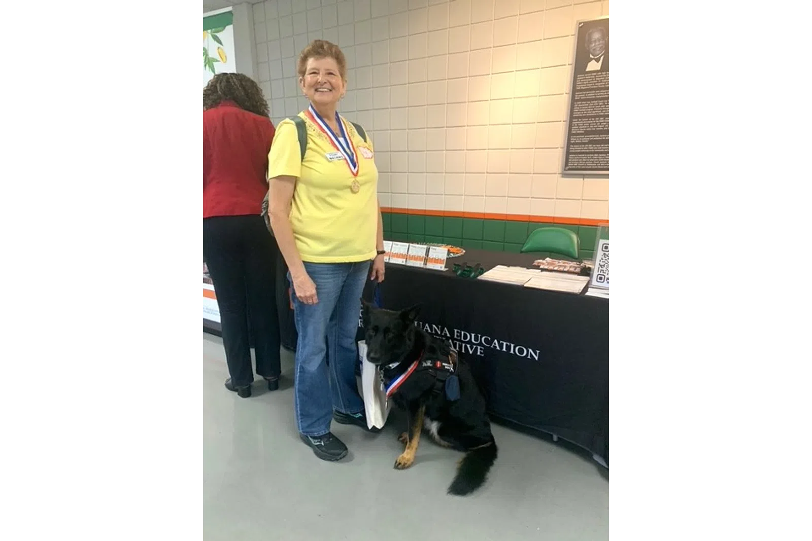 Woman in yellow shirt standing next to a German Shepherd service dog at an education event booth.