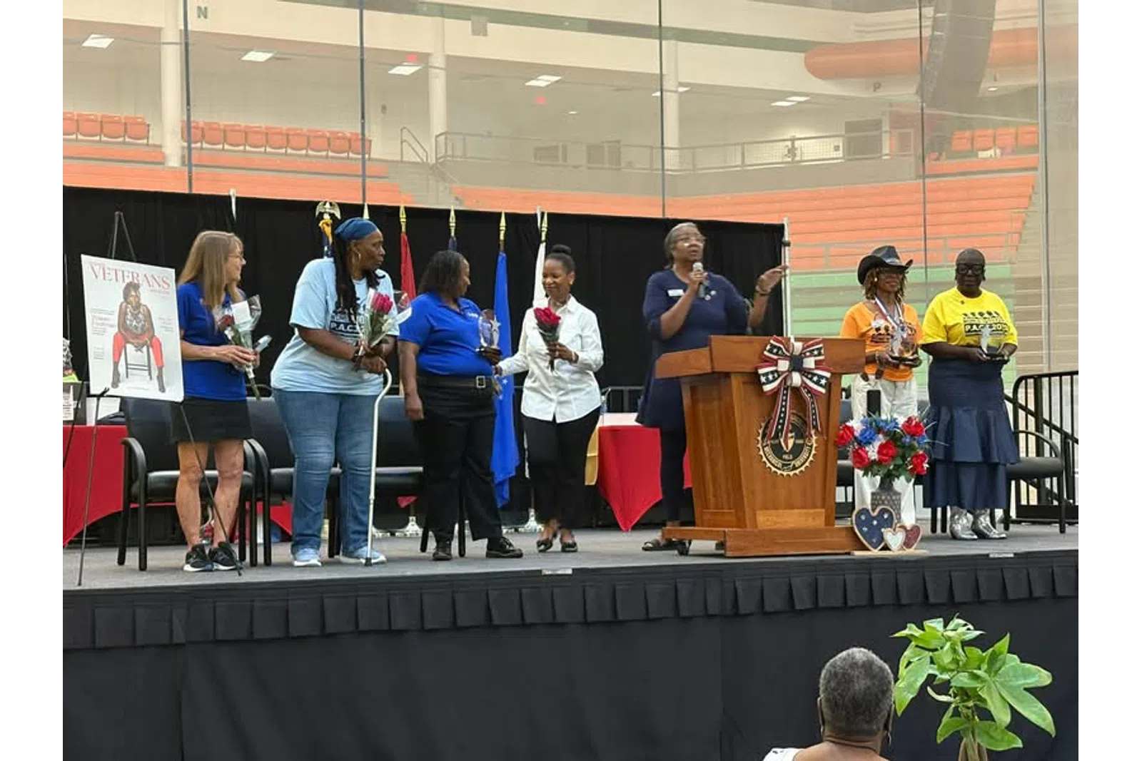 Group of women on stage holding flowers and awards during a veterans appreciation event in a gymnasium.