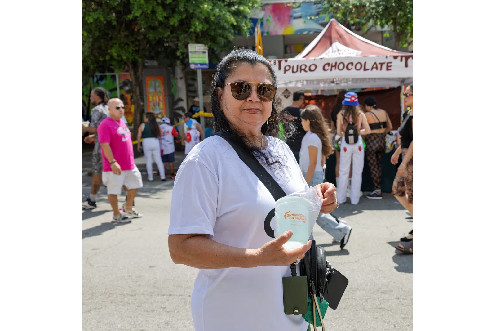Woman wearing sunglasses and white shirt holding a cup at an outdoor festival with a Puro Chocolate tent in the background.