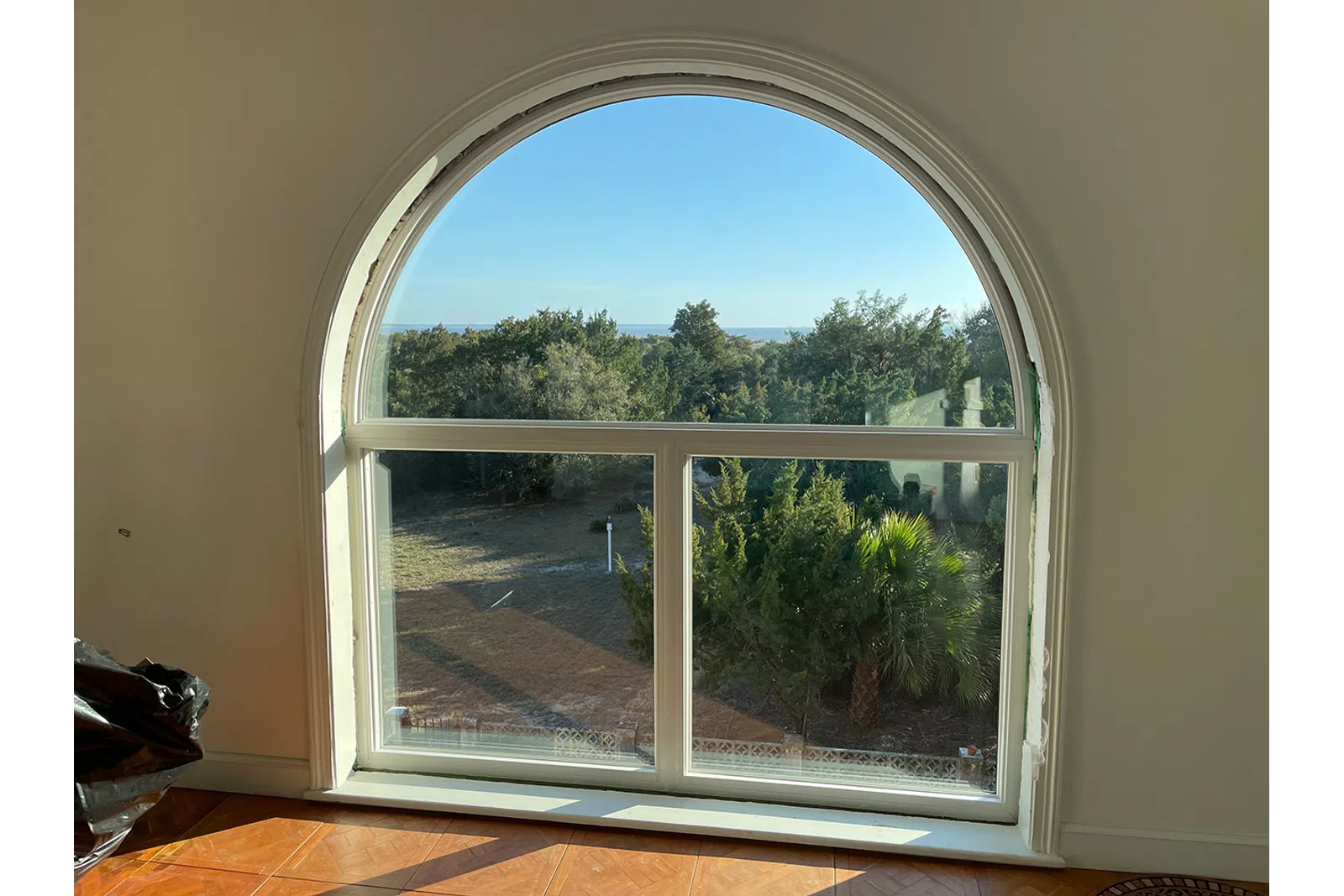 Arched window with white frame overlooking trees and blue sky with sunlight casting shadows on hardwood floor.