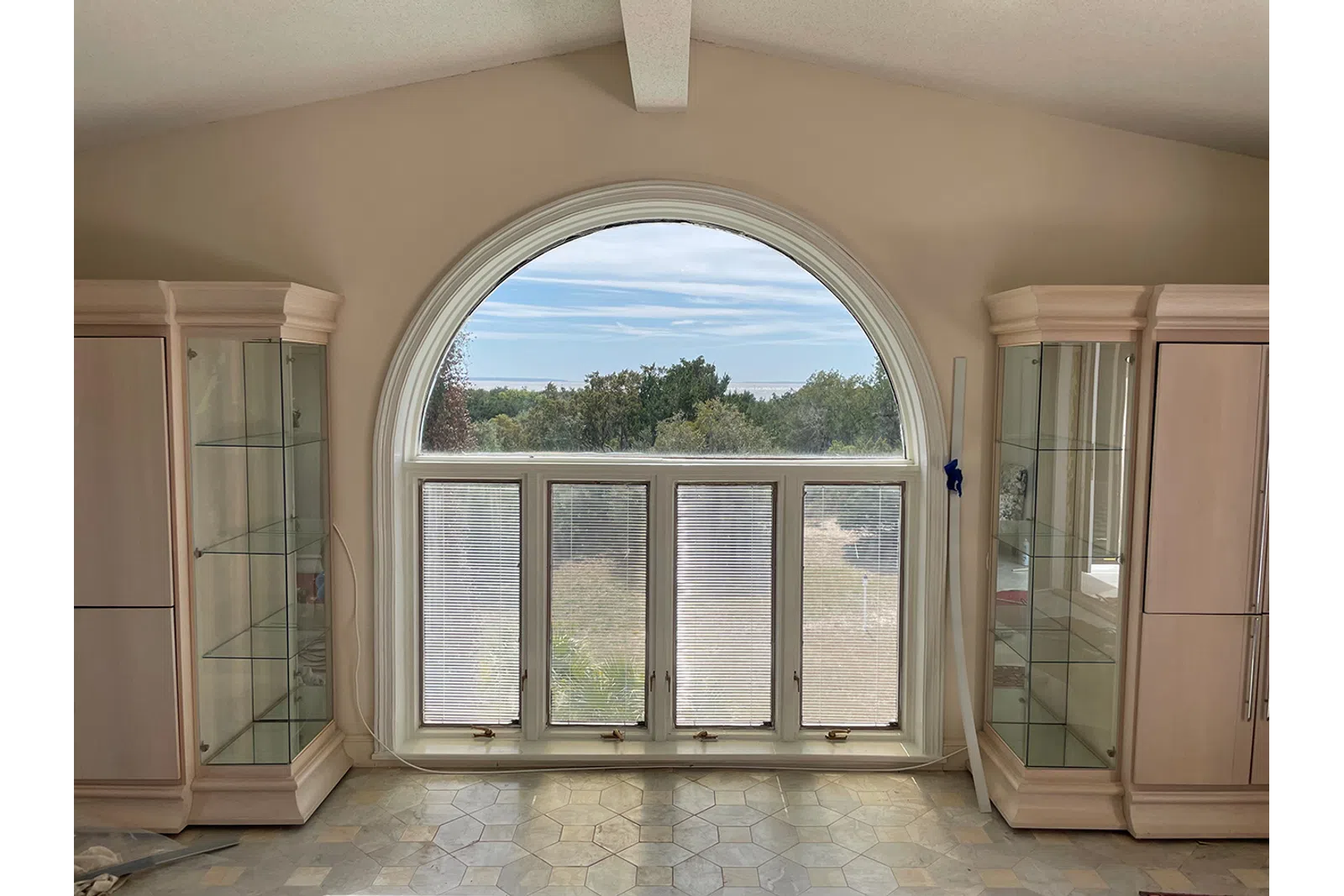 Bright room with large arched window, beige cabinets, glowing tiled floor, and outdoor greenery view.