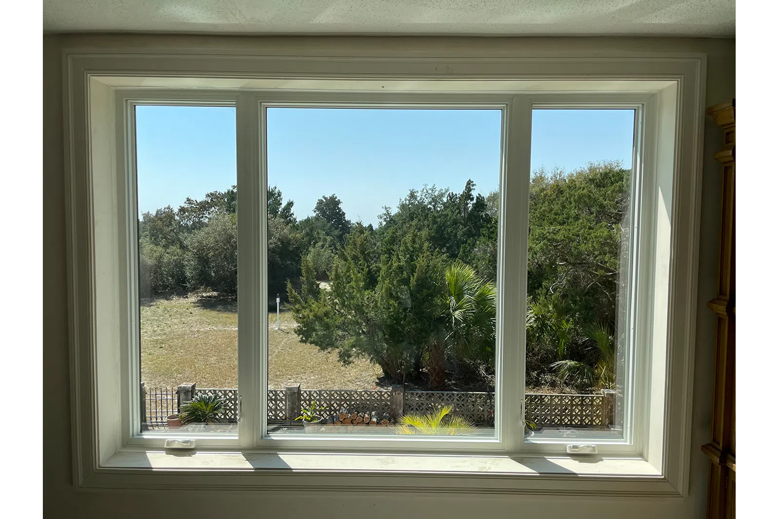 View through a large three-panel window showing trees, bushes, and a clear blue sky outside a fenced yard.