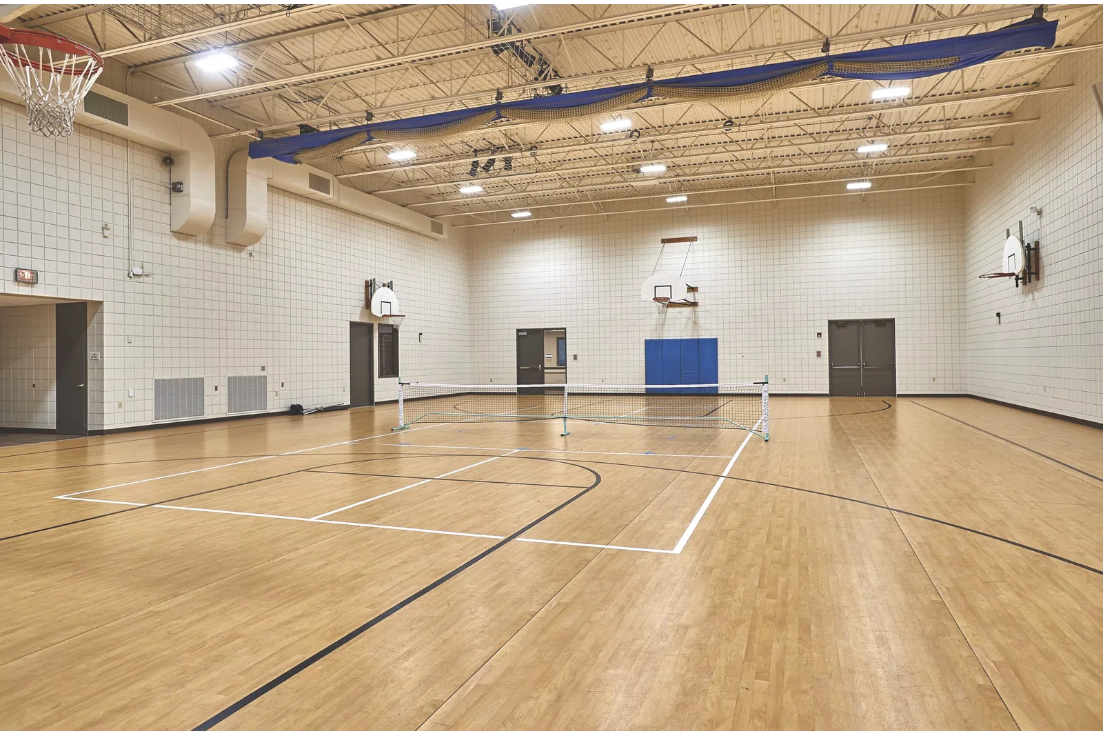 An empty indoor gym with a tennis court, basketball hoops, and wooden flooring.