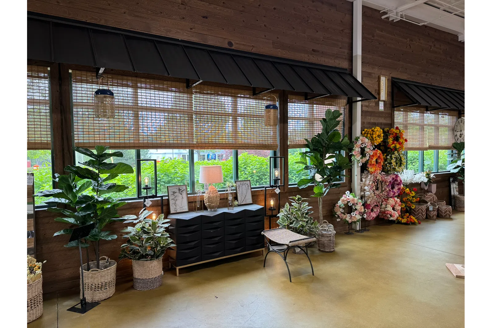 Indoor garden shop display with potted plants, woven baskets, floral arrangements, and a black storage cabinet under wooden windows.