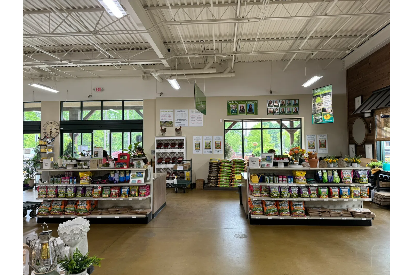 Interior of a bright gardening store with shelves of seeds, soil bags, gardening tools, and plants near large windows.