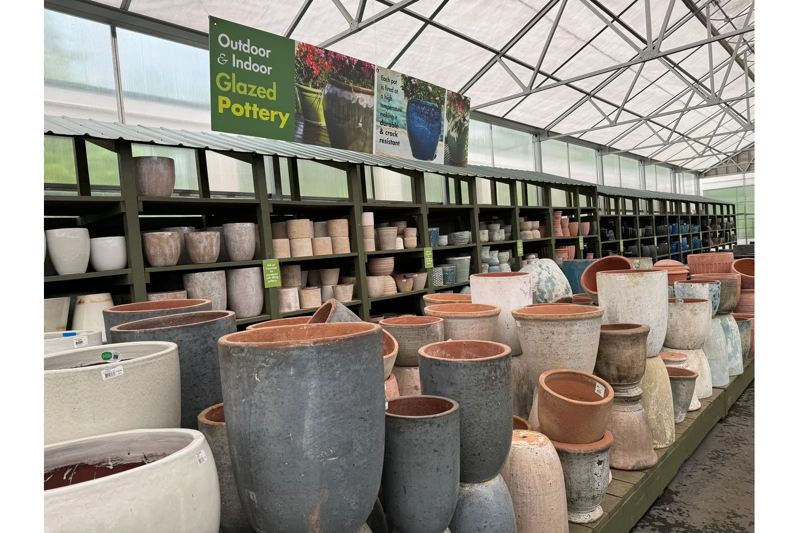 Rows of glazed pottery pots in various sizes displayed on shelves inside a greenhouse garden center.
