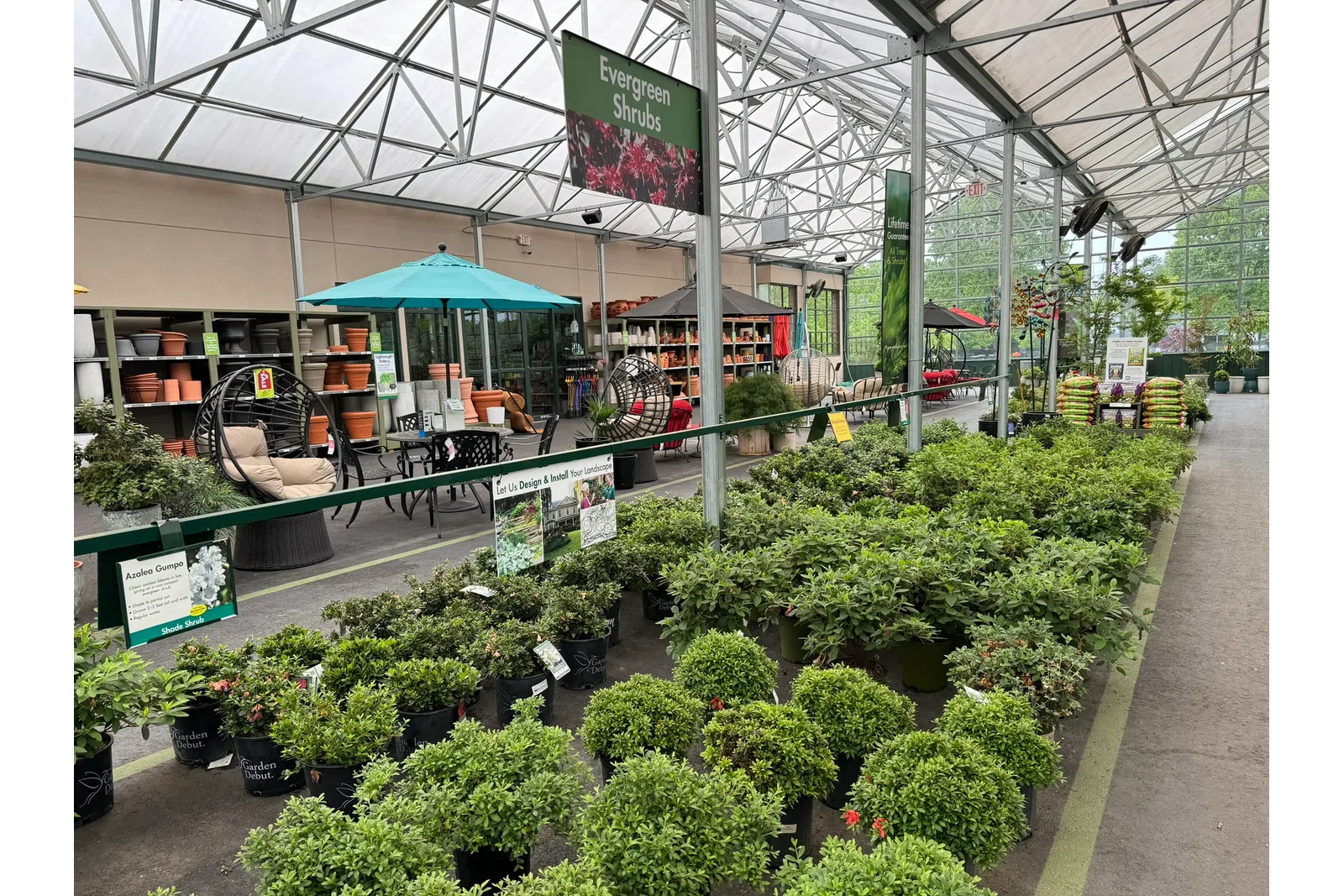 Indoor garden center with rows of evergreen shrubs, potted plants, patio furniture, and gardening supplies under a large greenhouse roof.