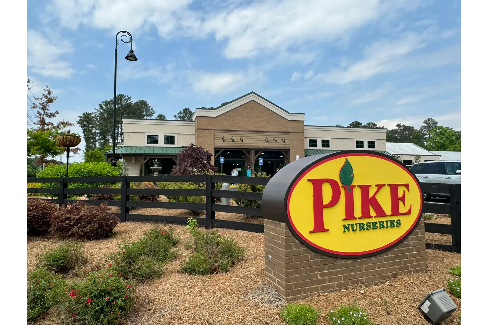 Pike Nurseries sign in front of a landscaped garden and a brick building under a partly cloudy sky.
