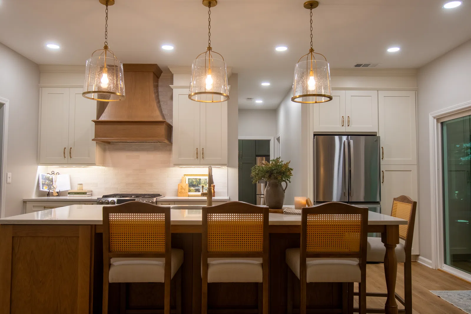 Modern kitchen with wooden island, rattan chairs, pendant lights, stainless steel fridge, and white cabinetry