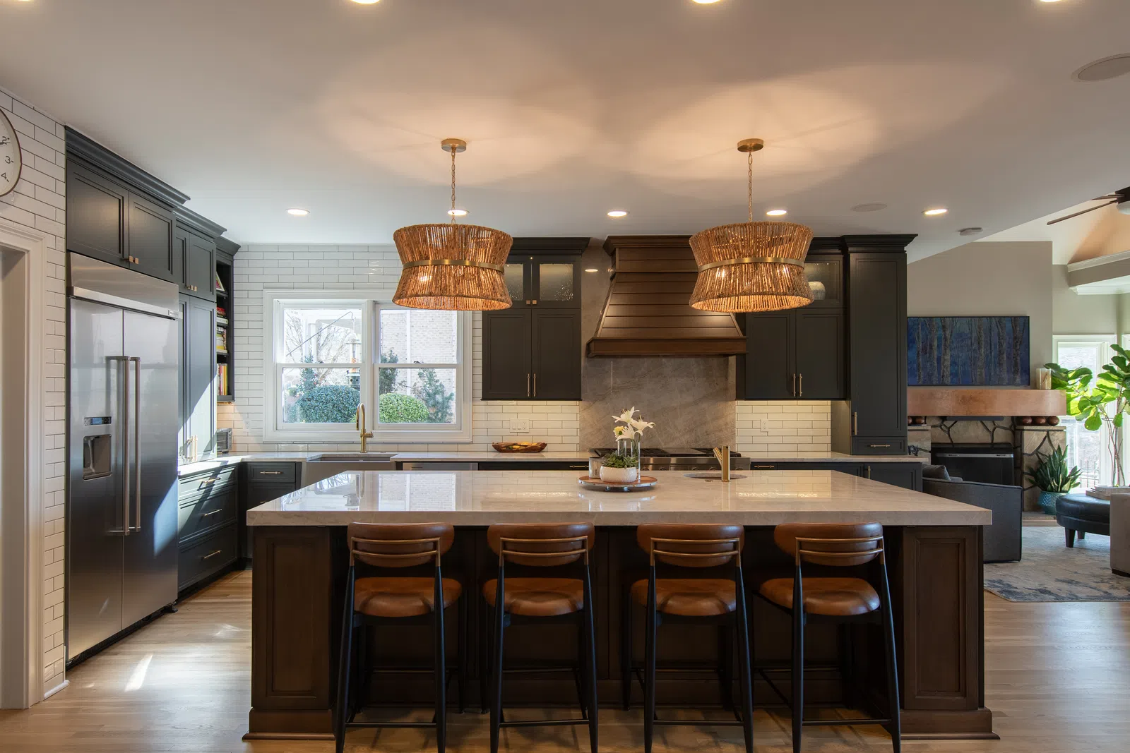 Modern kitchen with large marble island, dark cabinets, wicker pendant lights, and stainless steel appliances.