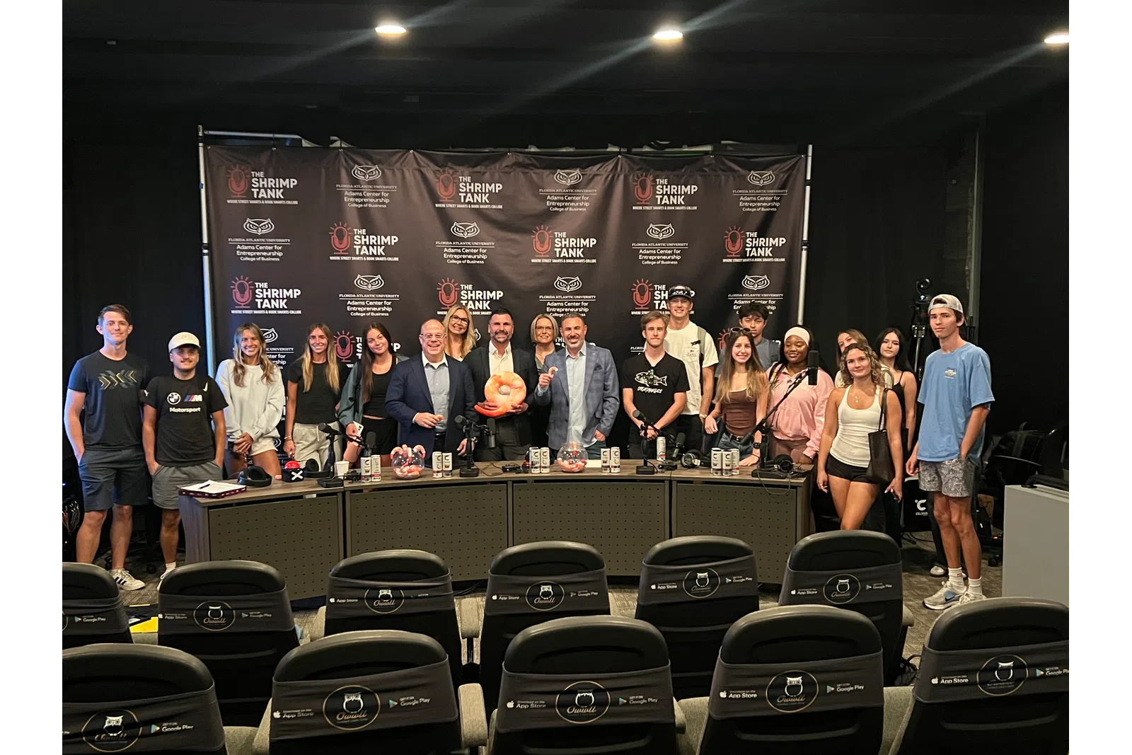Group of people posing in front of The Shrimp Tank backdrop at entrepreneurship event with a panel table and prizes.