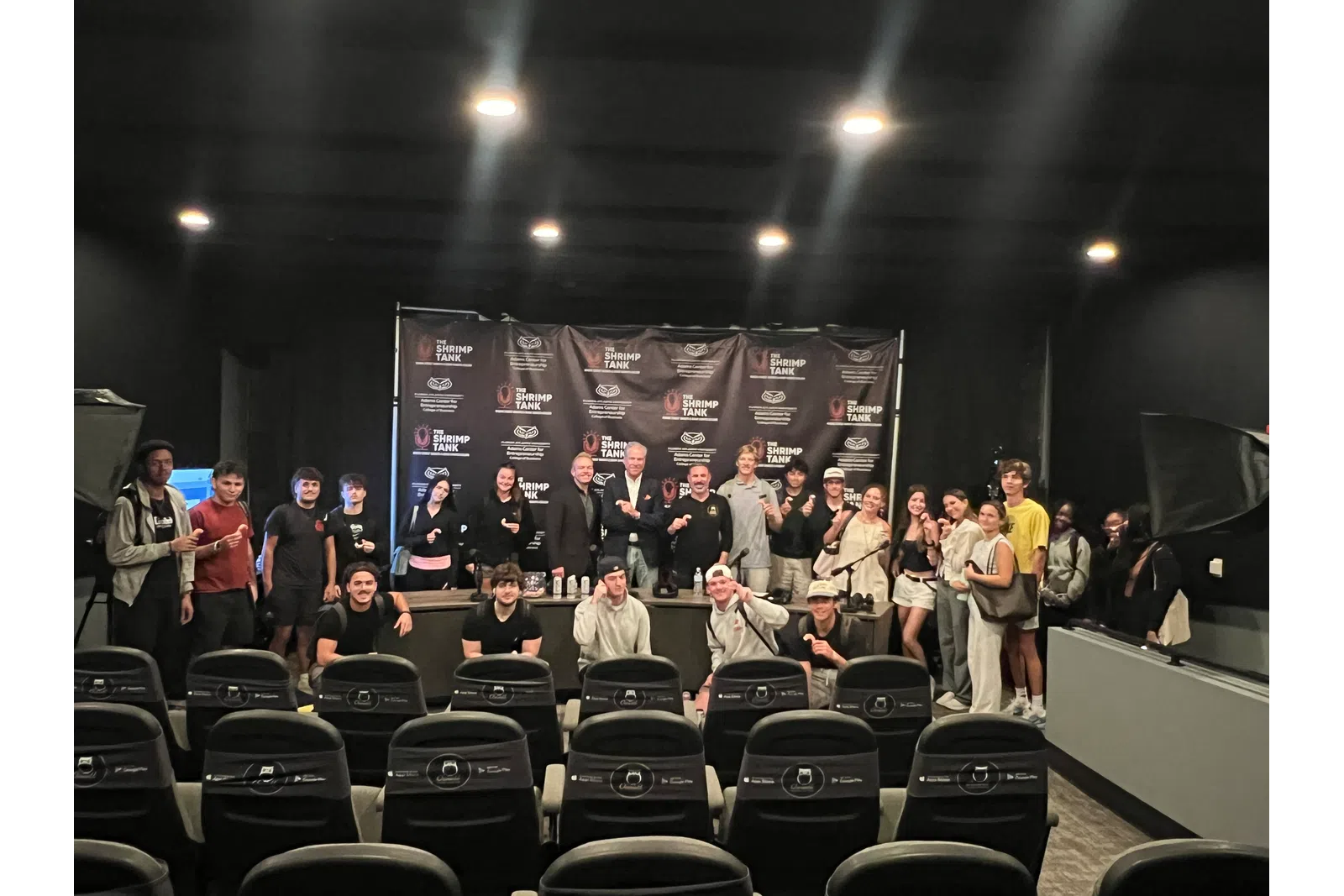 Group of people posing in a theater with Shrimp Tank branded backdrop and empty chairs in the foreground.