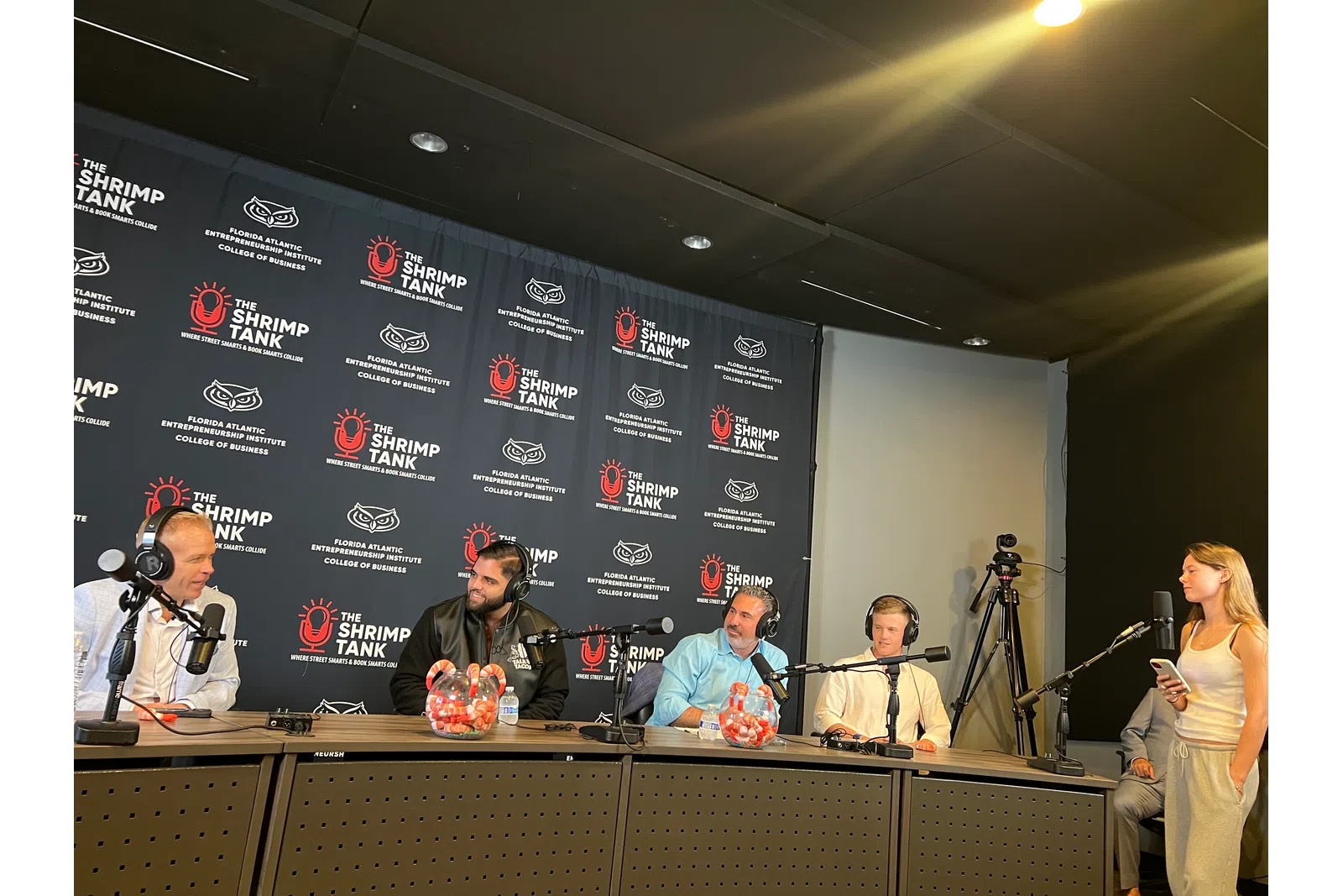 Panel of five people in a studio podcast setup with microphones, headphones, and branding backdrop for The Shrimp Tank.