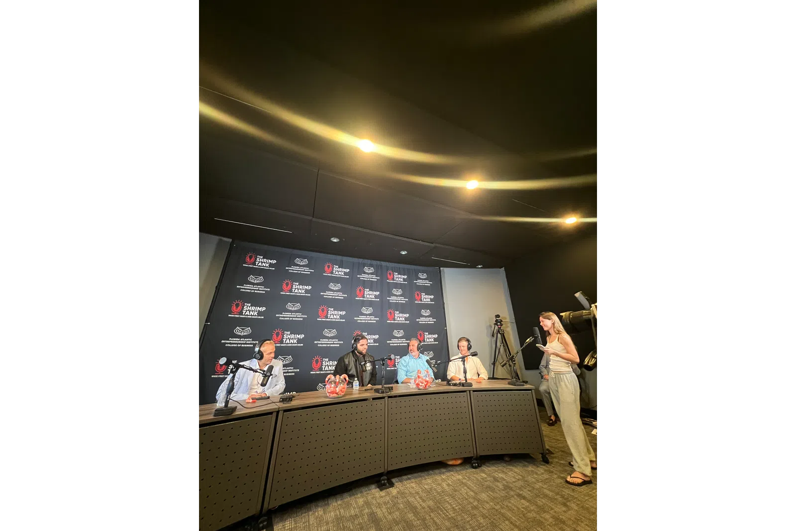 Panel discussion at The Shrimp Tank event with four speakers sitting behind a desk and a woman standing with a microphone.