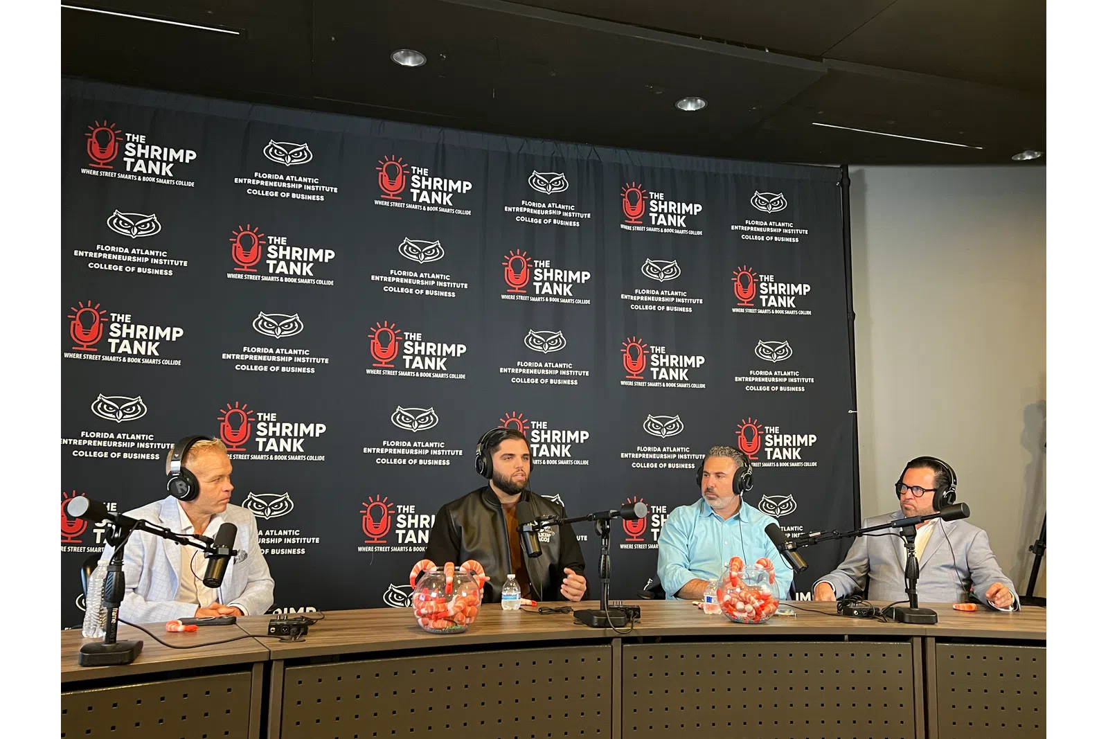 Four men wearing headphones talk into microphones at a table during The Shrimp Tank podcast recording.