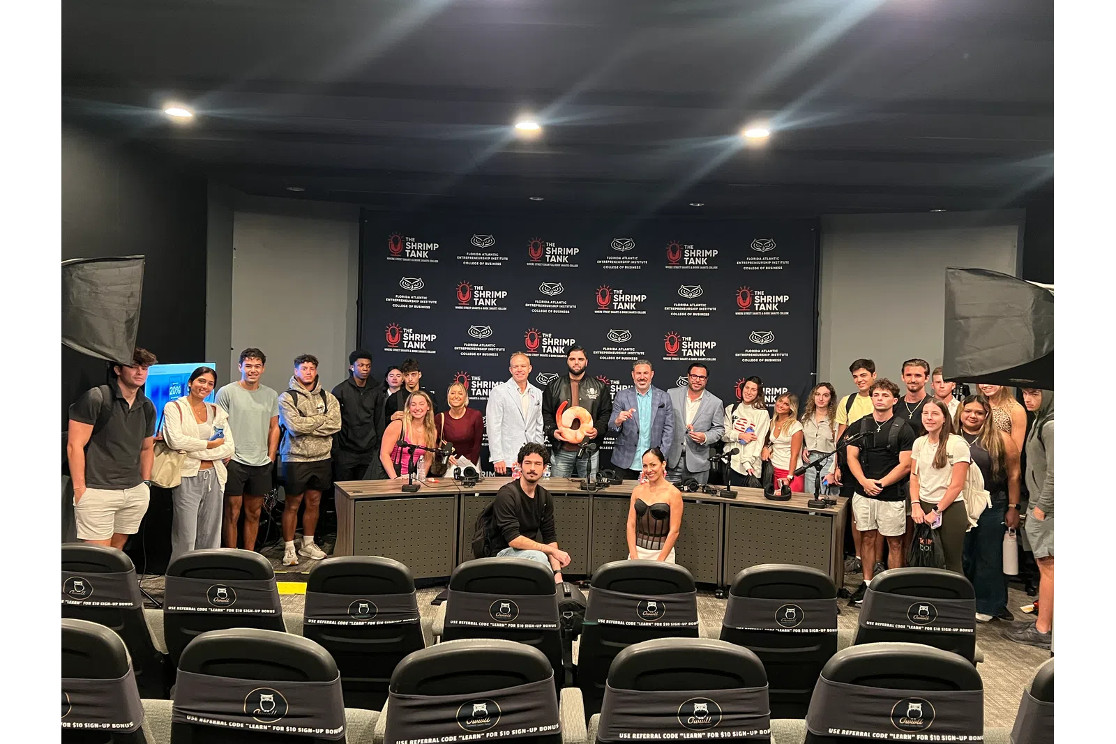 Group photo of diverse people standing behind a conference table with Shrimp Tank branding in a studio setting.