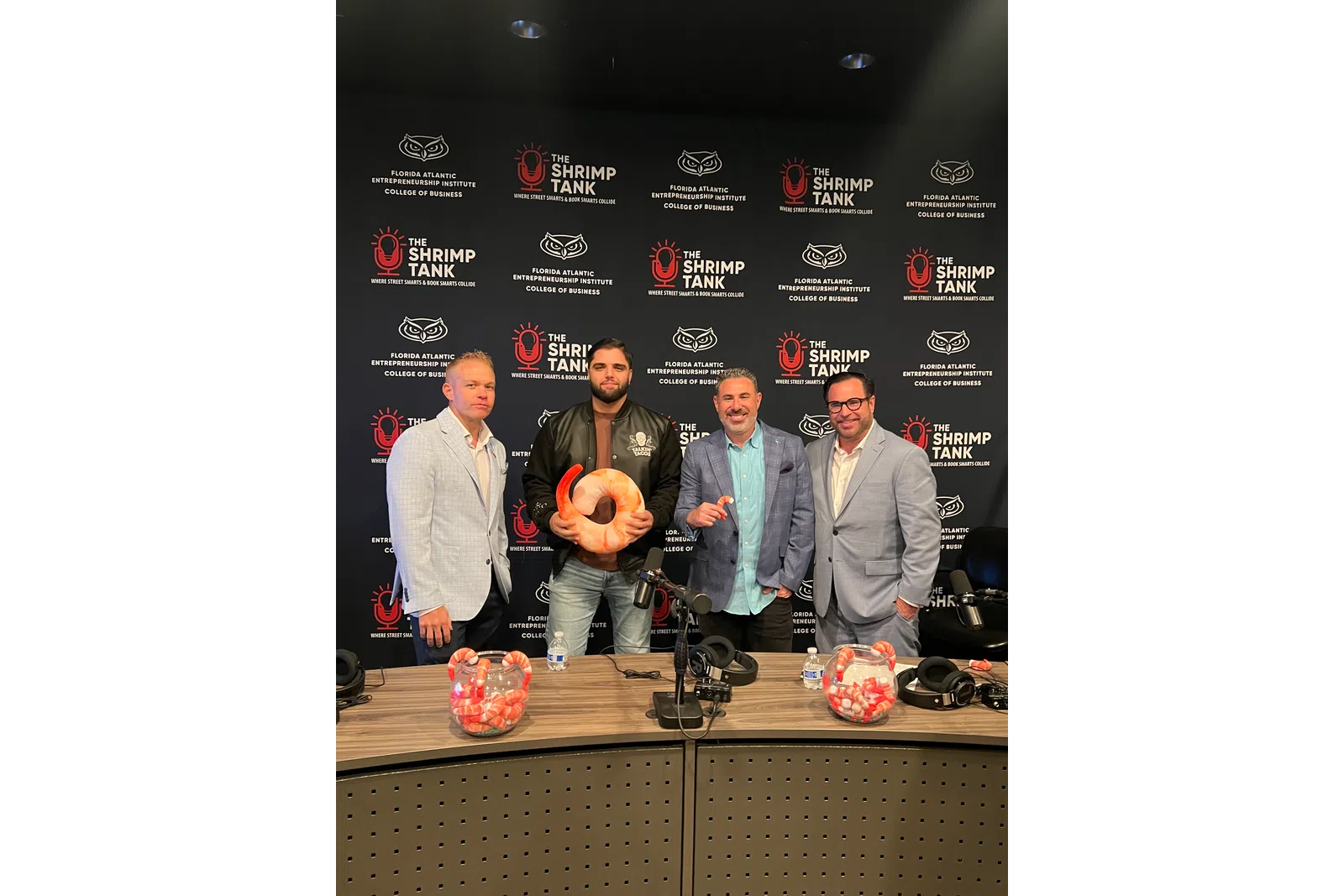 Four men standing in front of The Shrimp Tank and Florida Atlantic Entrepreneurship Institute backdrop, holding shrimp prop, podcast setup