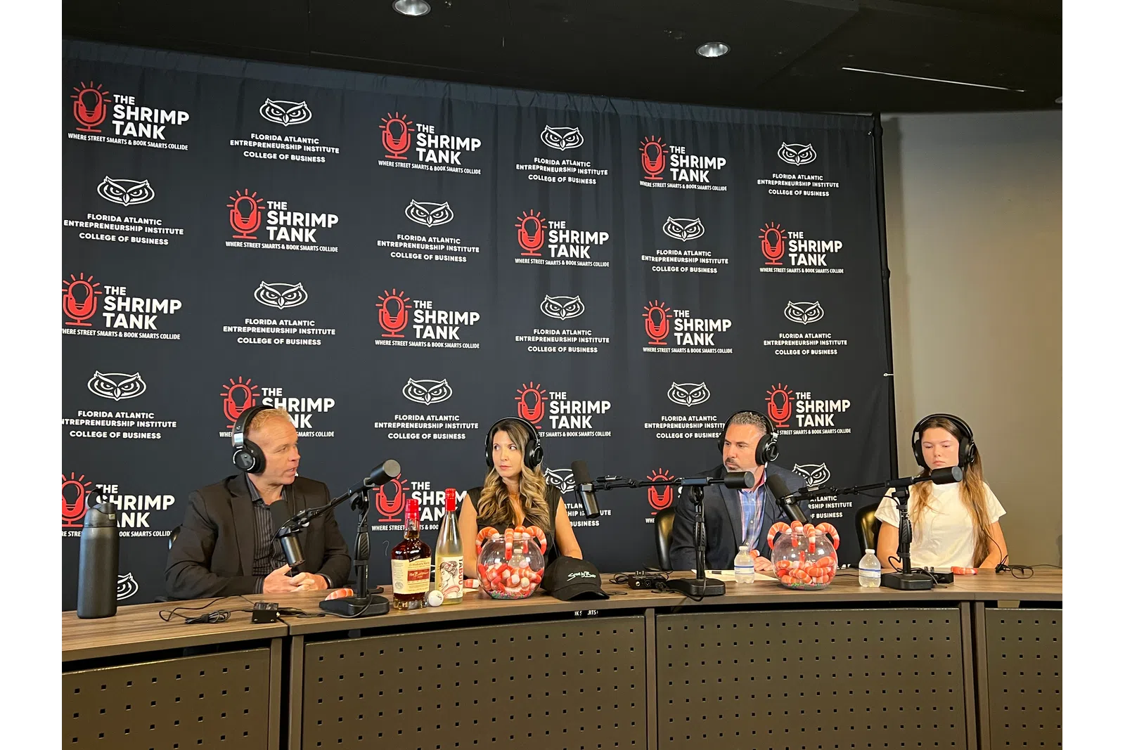 Four hosts wearing headphones seated at a podcast table with microphones and branded backdrop reading The Shrimp Tank