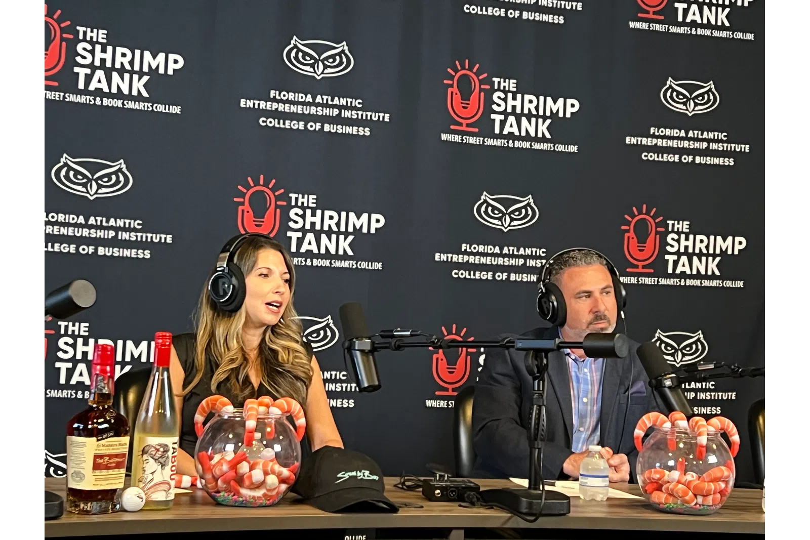 Two hosts wearing headphones speaking into microphones with branded backdrop and shrimp-themed props on table
