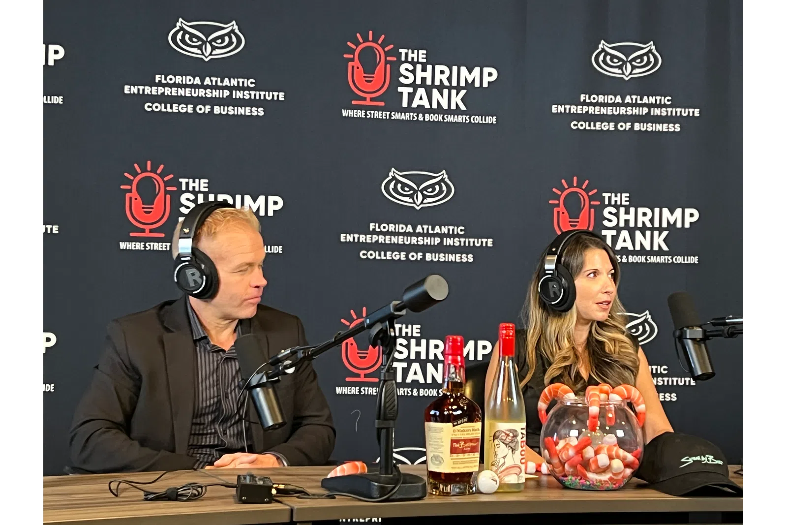 Two podcasters wearing headphones speak into microphones with branded backdrop and assorted drinks on table.