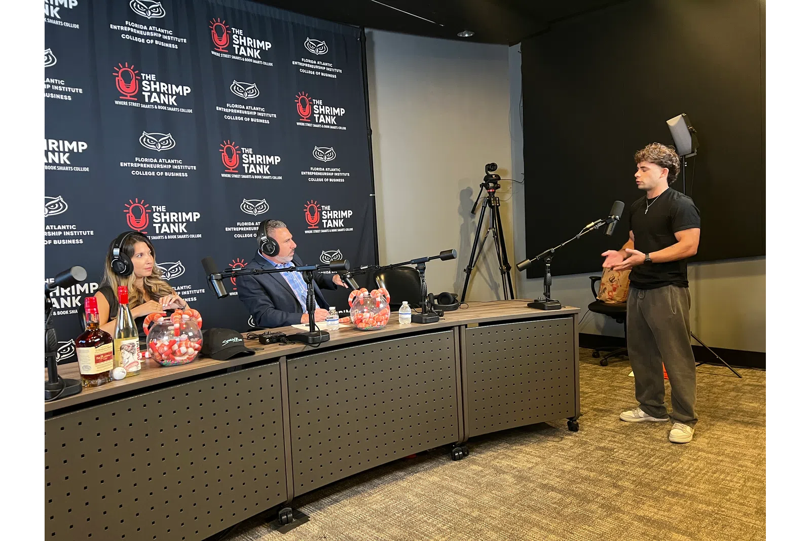 A man pitches to two judges seated at a table with microphones and branded backdrop for The Shrimp Tank podcast.