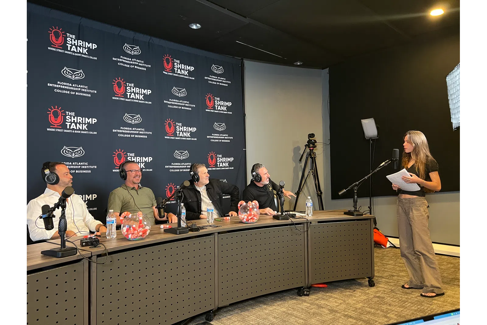 Panel of four men with headsets at a desk facing a woman standing with papers in a podcast studio with branded backdrop.