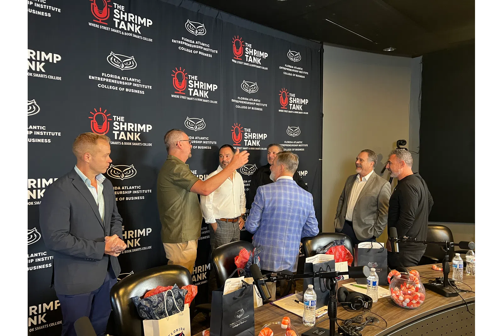 Group of six men in business attire talking at The Shrimp Tank event with overlaid logo and text backdrop.