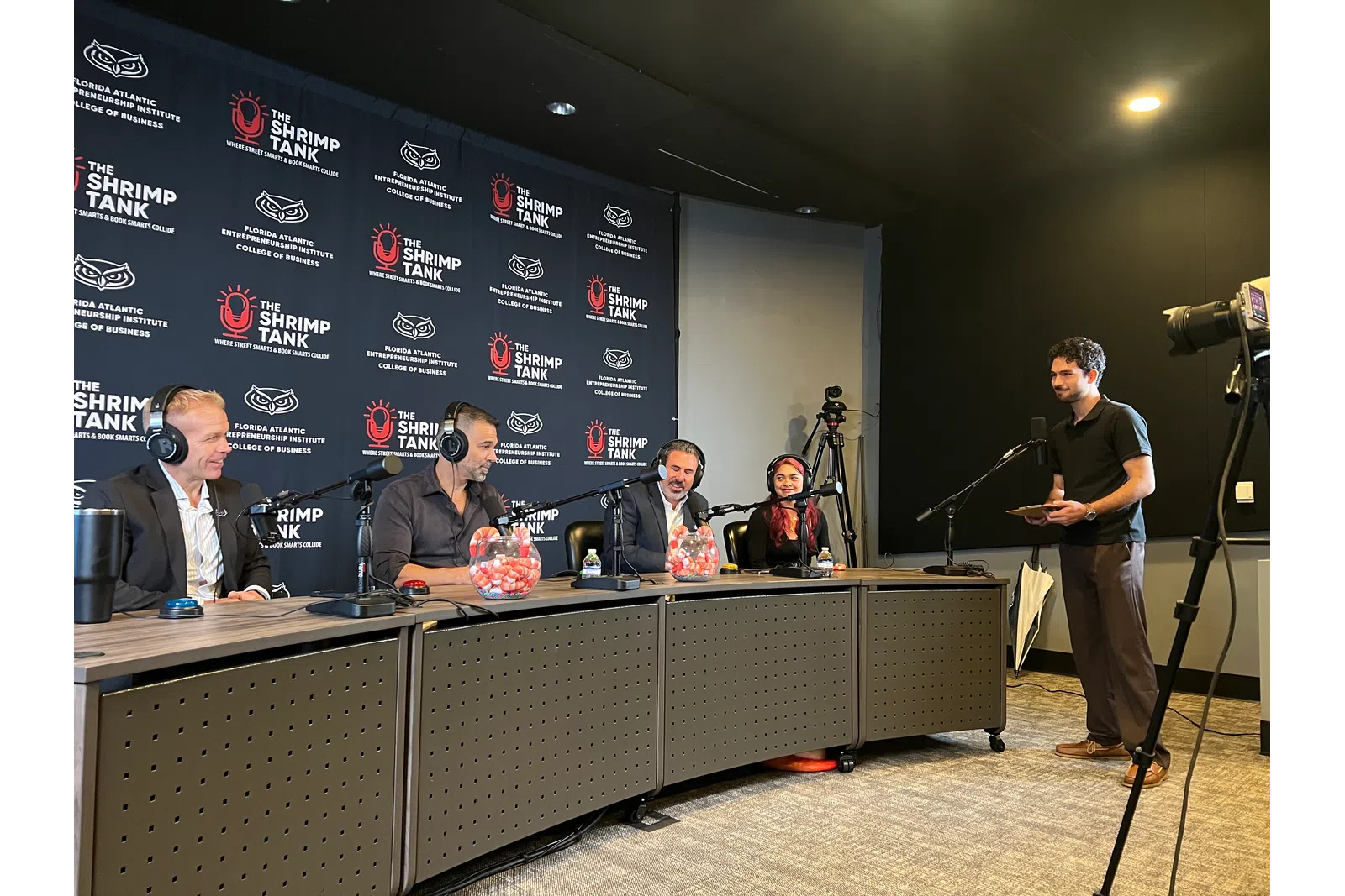 Panel of four judges in a podcast studio discussing on microphones with a host standing near a camera setup.