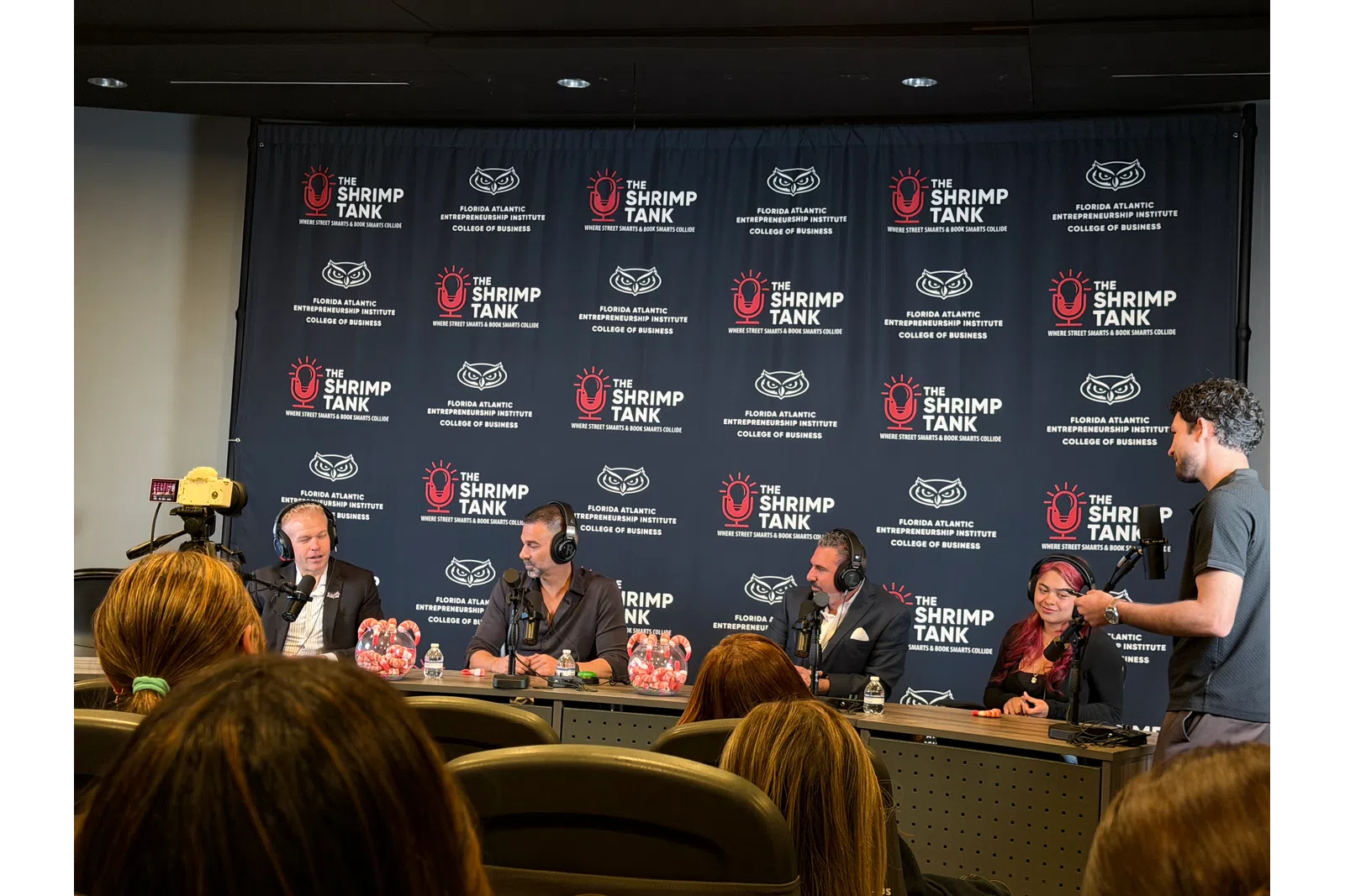 Panel of speakers at The Shrimp Tank event with branded backdrop and audience in foreground