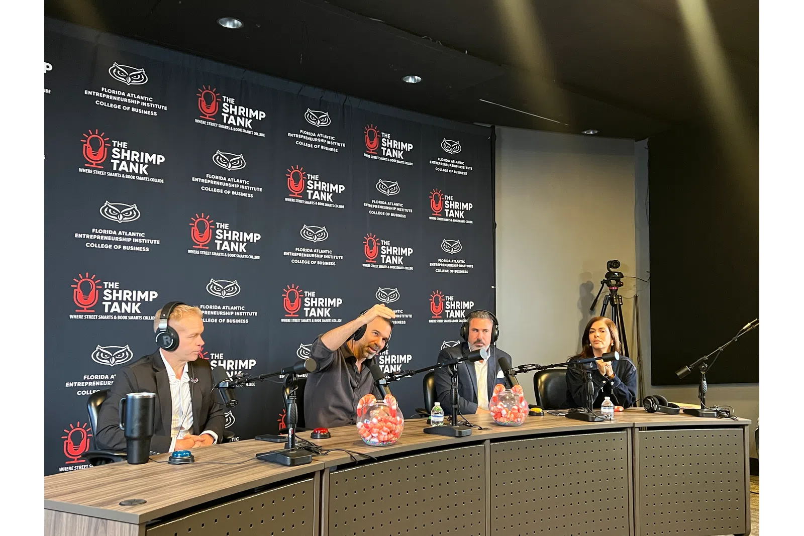 Panelists wearing headphones participate in a podcast recording at The Shrimp Tank event with branded backdrop.