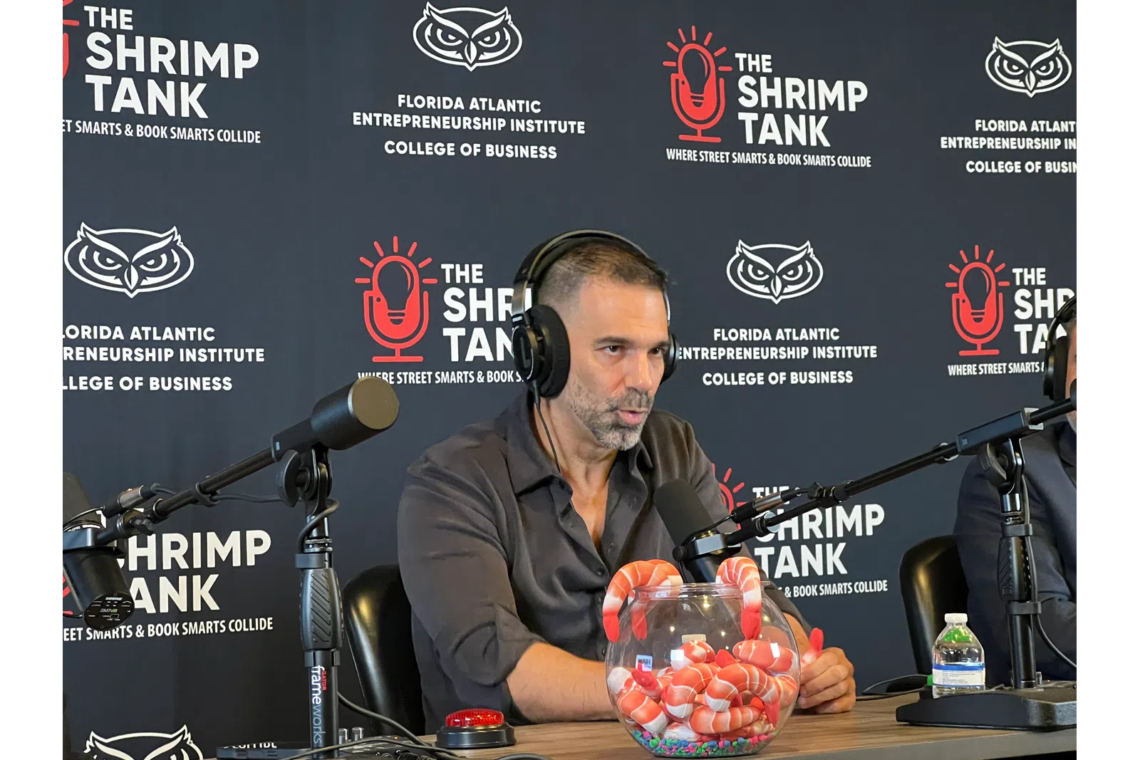 Man with headphones speaking into microphone at The Shrimp Tank event with branded backdrop and shrimp bowl