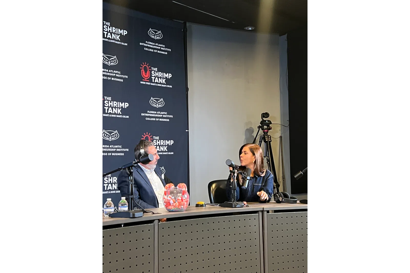 Two people recording a podcast episode at a table with microphones and a backdrop for The Shrimp Tank show.