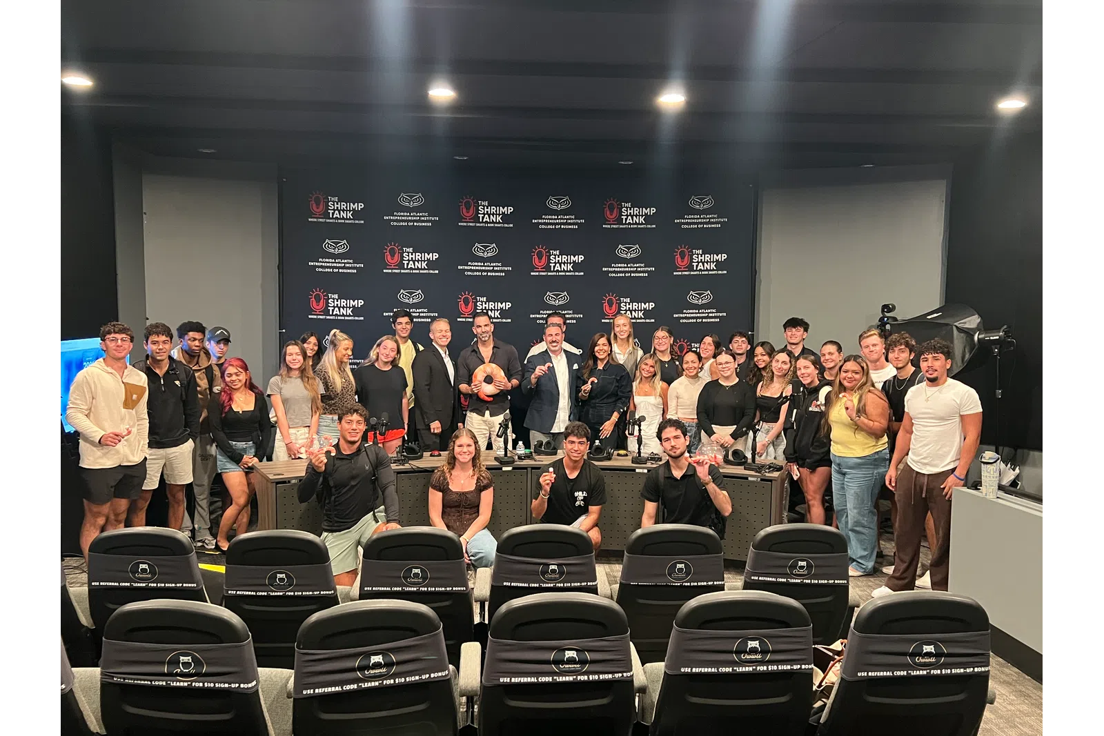 Group photo of diverse young adults and presenters at The Shrimp Tank event in a studio with branded backdrop and seating.