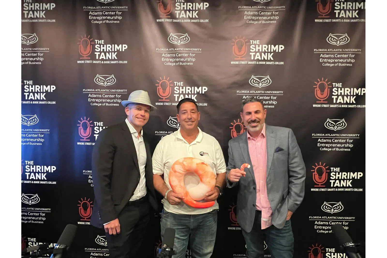 Three men posing in front of a Shrimp Tank backdrop, one holding a large shrimp prop, at an entrepreneurship event.
