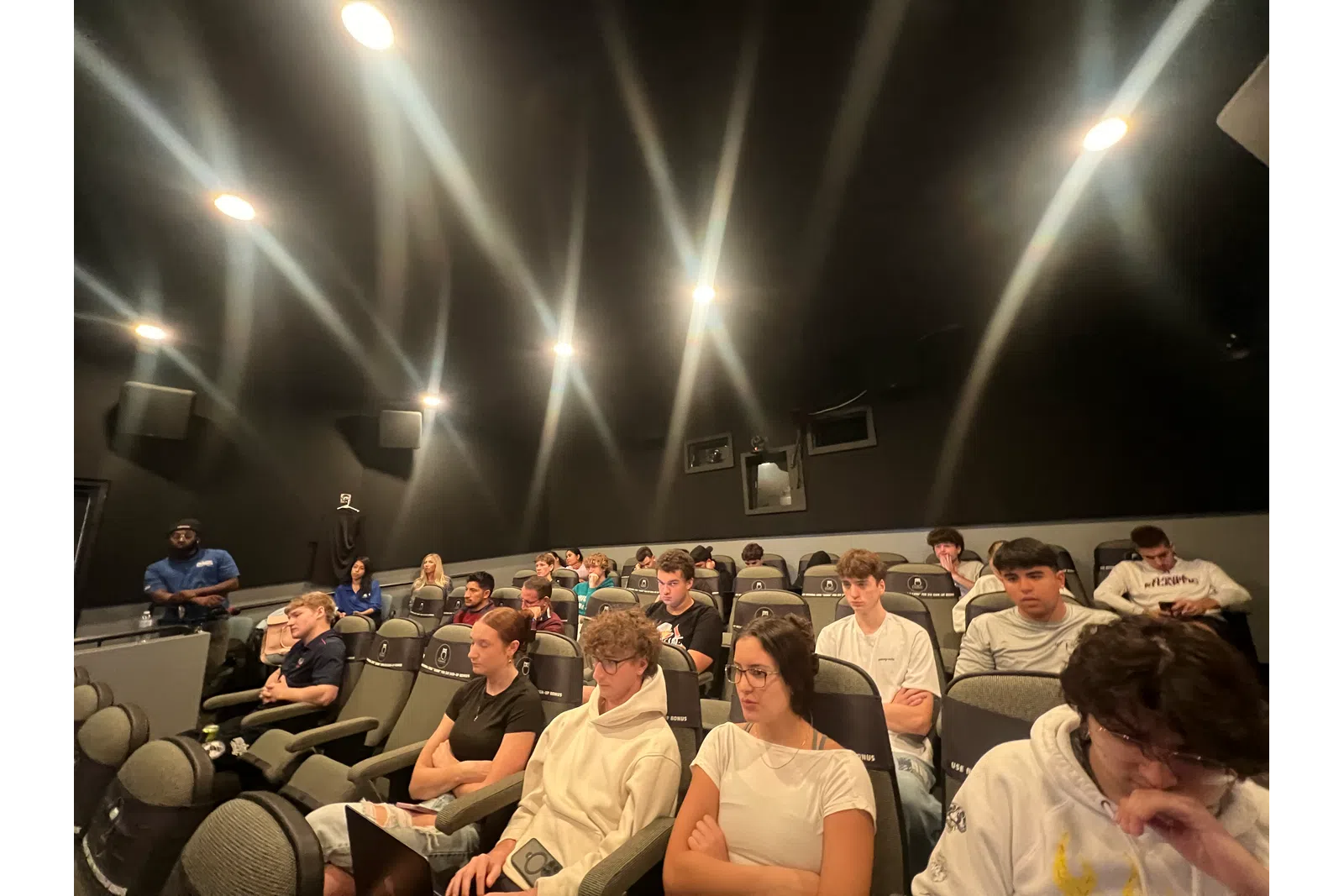 Group of young adults seated attentively in a dimly lit theater or lecture room with black walls and bright ceiling lights.