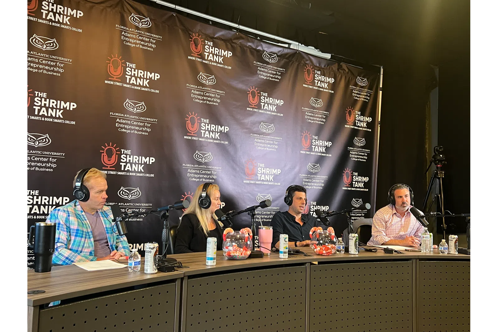 Four hosts with microphones and headphones recording a podcast at The Shrimp Tank event table with branded backdrop.