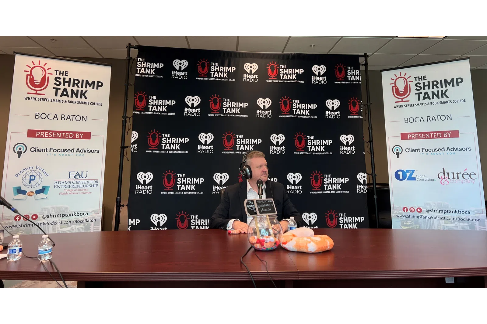 a person sitting at a table with a microphone and a sign behind him