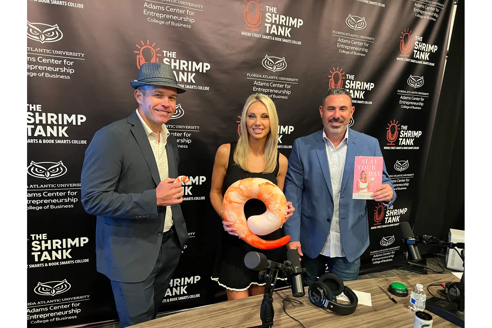 Three people posing with a shrimp-shaped pillow and a book in front of The Shrimp Tank event backdrop at Florida Atlantic University.