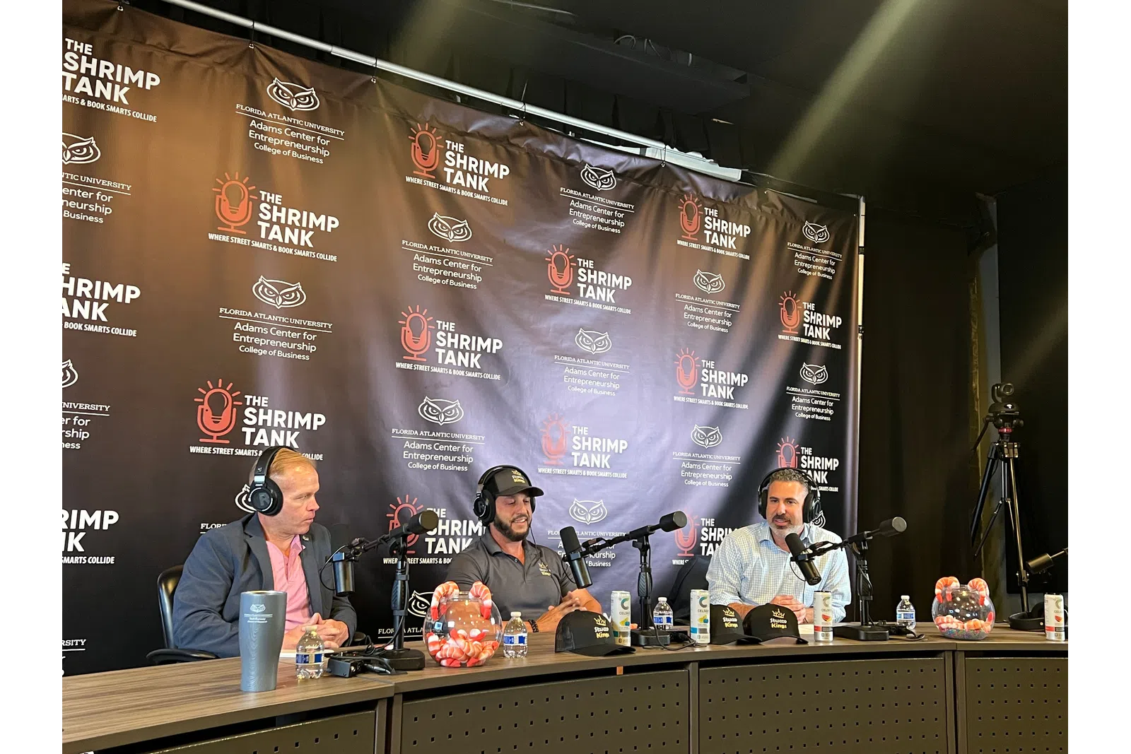 Three men with headsets and microphones seated at a table with The Shrimp Tank branded backdrop during a podcast or interview.