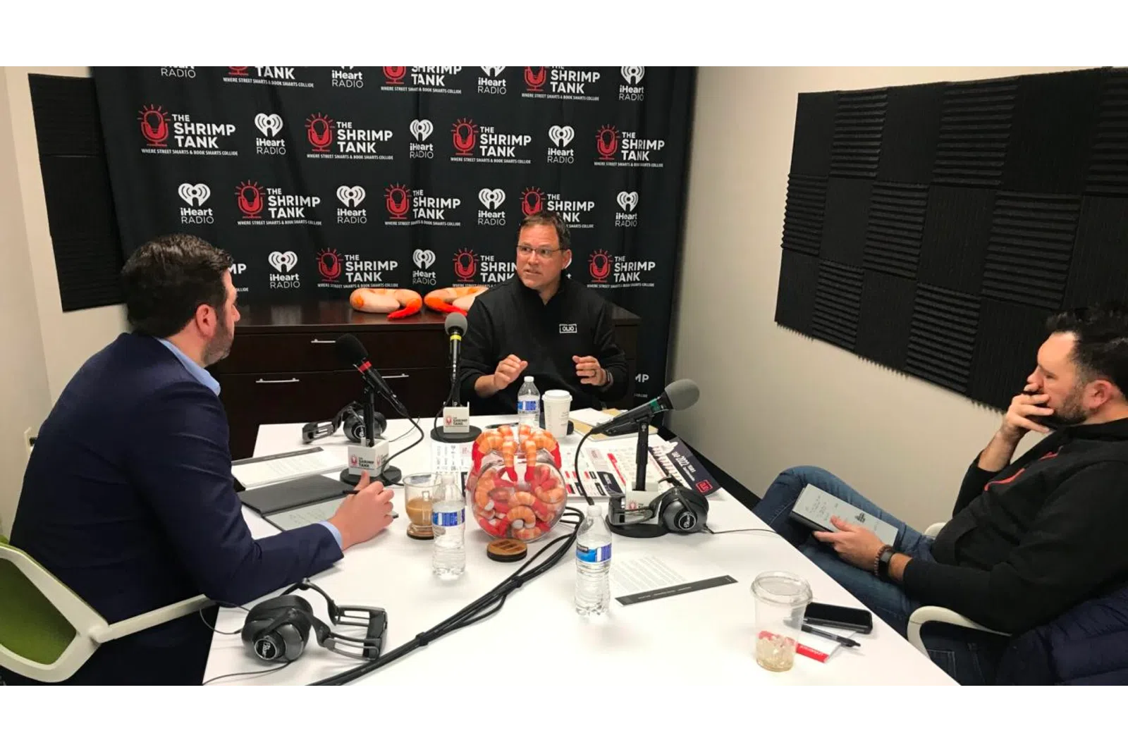 a group of men sitting at a table with microphones and a blackboard