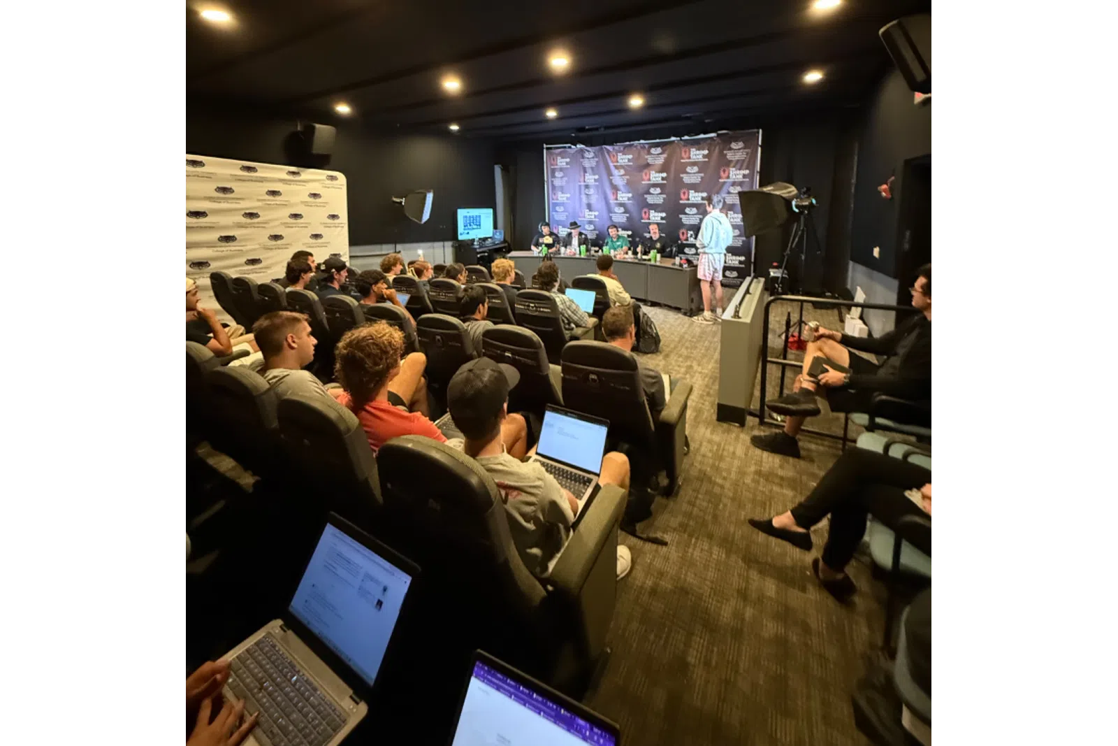 Audience attending a podcast panel discussion in a small theater room with laptops and microphones setup