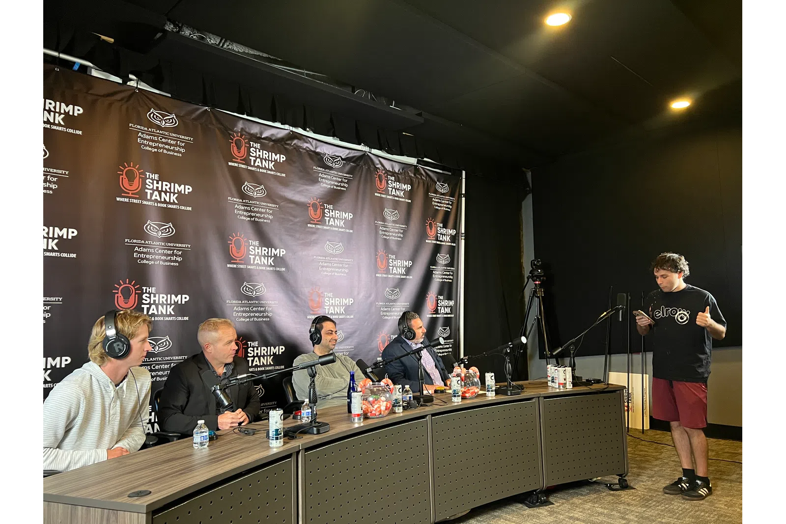 Four judges seated at a table with microphones listening to a standing contestant during The Shrimp Tank pitch event.