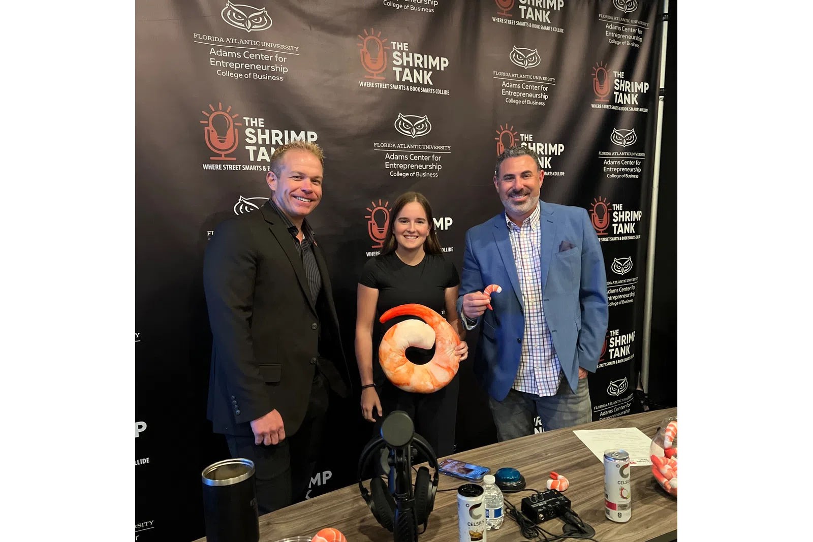 Three people posing at a Shrimp Tank event backdrop, one holding a large shrimp plush, with drinks and microphones nearby.