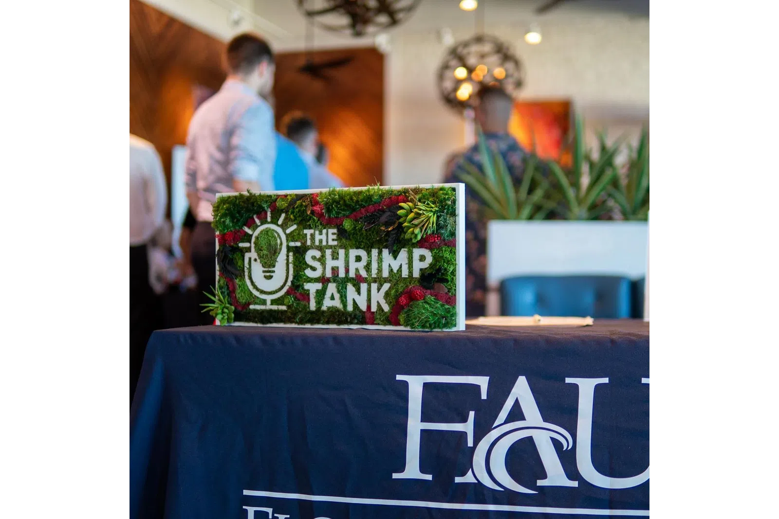 Decorative sign for The Shrimp Tank event on a table with FAU branding and blurred people in the background