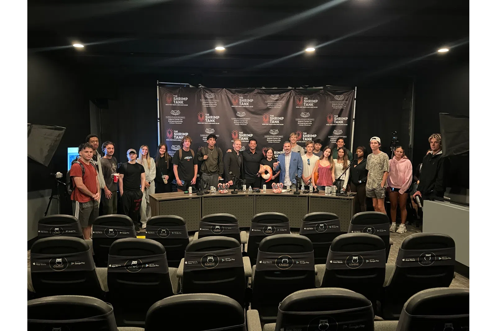 Group of people posing in front of a branded backdrop with chairs in the foreground under indoor lighting.